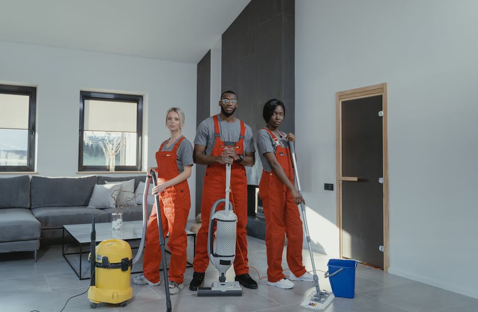 Three professional cleaners from Cleaners Richmond are engaged in surface cleaning within a modern living room, featuring a light-colored tiled floor, a grey fabric sofa, and large windows allowing na